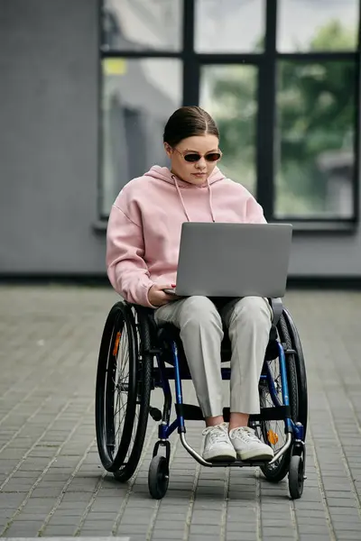 Stock image A young woman wearing sunglasses and a pink hoodie sits in a wheelchair outside while using a laptop.