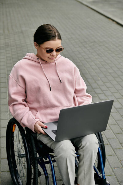 Молодая женщина в розовой толстовке сидит в инвалидном кресле на асфальтированном тротуаре, используя ноутбук.