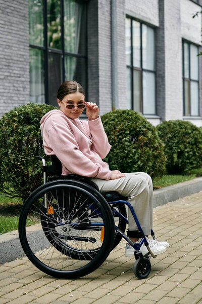 A young woman in a pink hoodie sits in a wheelchair on a sidewalk, adjusting her sunglasses with a confident expression.