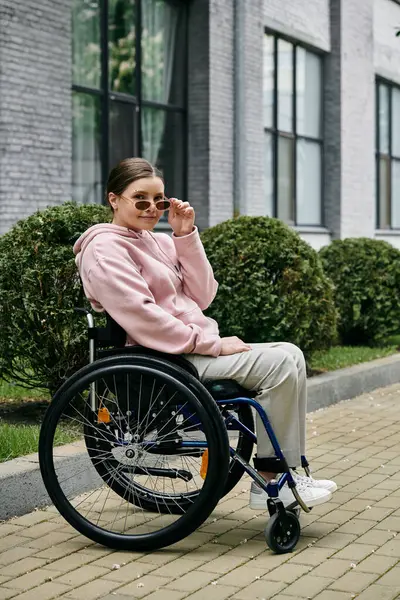 Pembe kapüşonlu genç bir kadın dışarıda tekerlekli sandalyede oturuyor, gülümsüyor ve güneş gözlüğünü ayarlıyor..
