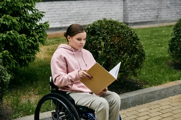 stock image A young woman in a pink hoodie sits in a wheelchair outside, reading a book.