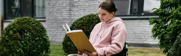 stock image A young woman in a pink hoodie sits in a wheelchair and reads a book outdoors.