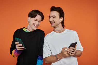 Canlı kıyafetli iki eşcinsel arkadaş telefonlarına bakarken turuncu bir arka planda gülüyorlar..