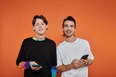 Şık ve canlı giyinmiş iki eşcinsel arkadaş turuncu bir arka plan önünde kameraya gülümsüyor..