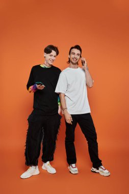 İki eşcinsel arkadaş, eşsiz moda anlayışlarını sergileyerek, turuncu bir arka plana şık bir şekilde poz veriyorlar..