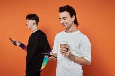 Parlak elbiseli iki şık genç, telefonlarına yapıştırılmış turuncu bir arka fona karşı birlikte gülümsüyorlar..