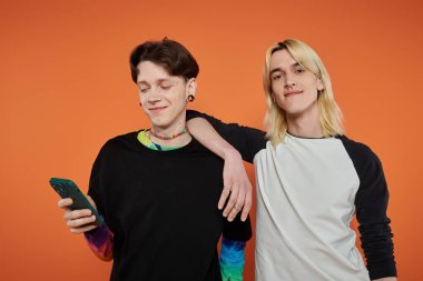 Renkli, şık giyinmiş iki genç arkadaş turuncu bir arkaplanda yan yana durur..