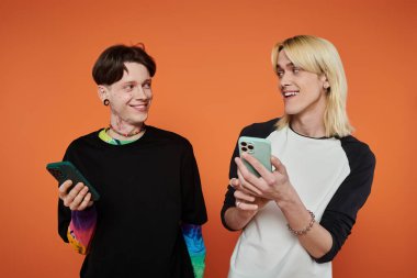 Parlak turuncu arka planda şık giyinmiş iki genç arkadaş gülümsüyor ve telefonlarına bakıyor..