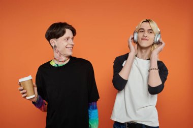 İki genç arkadaş, şık giyinmiş, turuncu arka planda duruyorlar, biri kahve tutuyor ve diğeri müzik dinliyor..