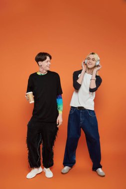 Şık ve renkli giyinmiş iki genç arkadaş turuncu bir arka planda dururlar..