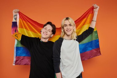 İki genç arkadaş, elinde gökkuşağı gururu bayrağıyla turuncu bir arkaplanın önünde gülümsüyor..