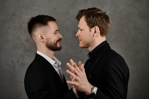 stock image A loving gay couple stands in front of a gray backdrop, both wearing elegant black suits.