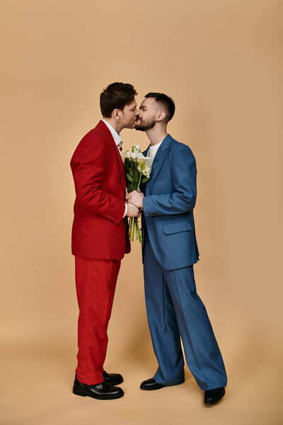 Двое мужчин в элегантных костюмах разделяют любящий поцелуй, держа букет цветов.