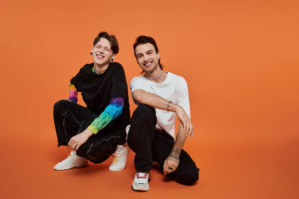 Two young queer friends in stylish outfits pose on a vibrant orange background.