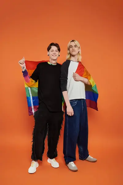 stock image Two young queer friends stand together, smiling and holding a rainbow pride flag against a vibrant orange backdrop.