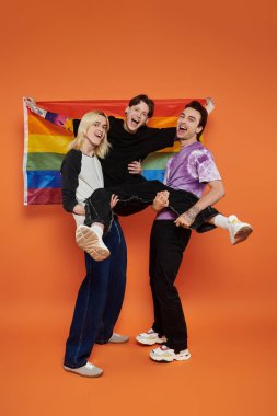 Three young friends in vibrant attire pose with a rainbow Pride flag against an orange background. clipart