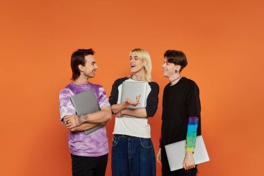 Turuncu arka planda şık giyinmiş, gülen ve dizüstü bilgisayar tutan üç eşcinsel arkadaş..