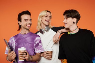 Canlı kıyafetler giyen üç genç arkadaş turuncu bir arka plana karşı birlikte poz veriyorlar..