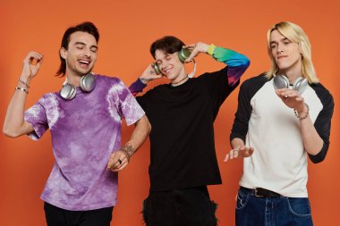 Şık giyinen, dans eden ve canlı turuncu arka planda poz veren üç eşcinsel arkadaş..