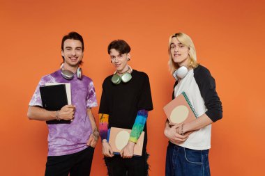 Eşcinsel topluluğundan üç genç, parlak turuncu bir arkaplana karşı şık ve canlı kıyafetler içinde poz veriyorlar..