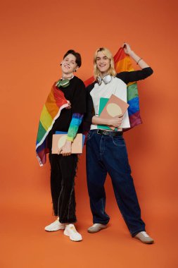 Omuzlarında gökkuşağı bayraklarıyla gururla duran iki eşcinsel arkadaş parlak turuncu bir arka plana karşı kitaplarını tutuyorlar..
