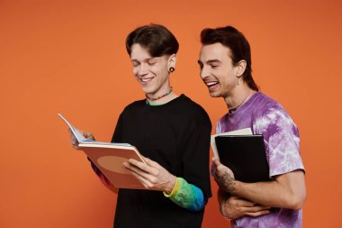 Canlı giyinmiş iki eşcinsel arkadaş turuncu bir arka planda duruyorlar, biri kitap okurken diğeri gülüyor..