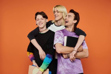 Üç eşcinsel arkadaş birlikte poz veriyor, gülümsüyor ve gülüyorlar, turuncu bir arkaplan önünde..