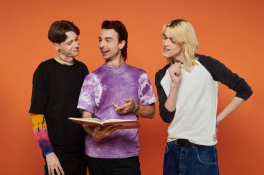 Tarz sahibi, renkli giyinmiş üç genç arkadaş turuncu bir arka plana yaslanmış, gülüyor ve bir kitabı paylaşıyorlar..