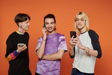 Renkli elbiseli üç genç arkadaş turuncu bir arkaplanda dikiliyorlar, her biri telefonlarıyla meşgul..