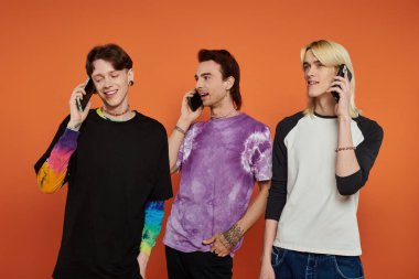 Şık ve canlı giyinmiş üç genç arkadaş, telefonlarıyla konuşurken parlak turuncu bir arkaplanda poz veriyorlar..