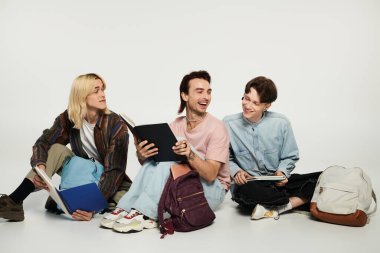 LGBTQIA topluluğunun bir parçası olan üç genç öğrenci gri bir arkaplanda oturuyor, gülüyor ve kitap paylaşıyorlar..