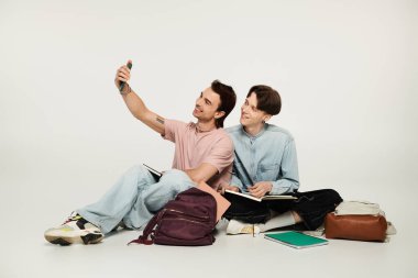 İki genç öğrenci gri bir arka planda oturup selfie çekiyorlar..