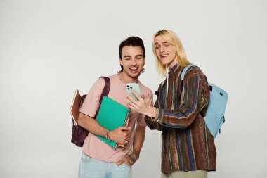 İki genç öğrenci, sıradan giyinmiş, gri bir arka plana karşı duruyor ve bir telefon ekranına bakarken gülüyorlar..