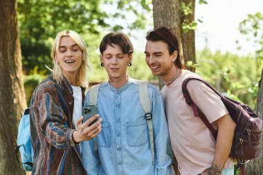 Sıradan giyinmiş üç genç arkadaş parkta dikilirken gülüyorlar ve telefona bakıyorlar..