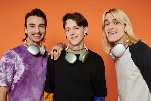 stock image Three young friends, wearing stylish attire, pose with headphones against a vibrant orange background.