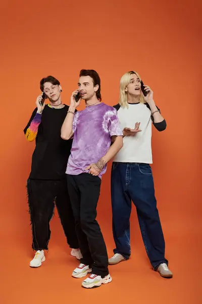 stock image Three queer friends in trendy attire, talking on cell phones against a bright orange background.