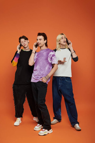 Three queer friends in trendy attire, talking on cell phones against a bright orange background.