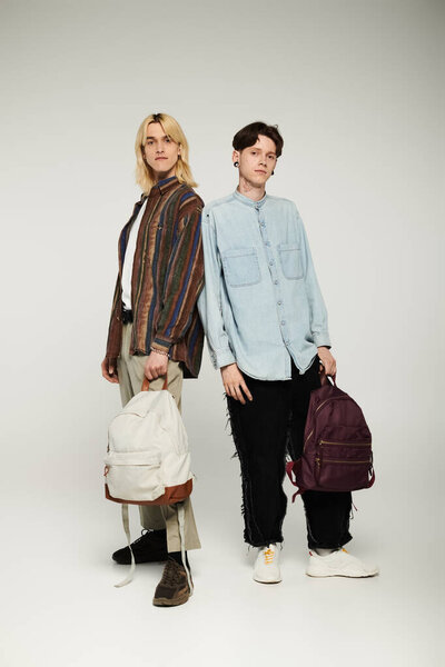 Two young queer students pose together in a studio, wearing casual attire and holding backpacks.
