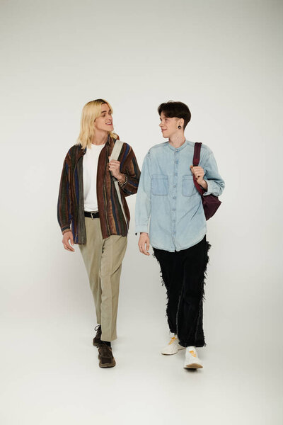 Two young students, part of the queer community, walk together on a grey background.