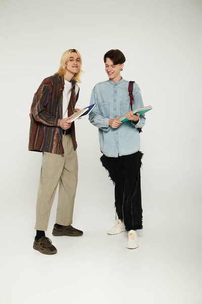 Two young queer students in casual attire hold books in front of a grey background.