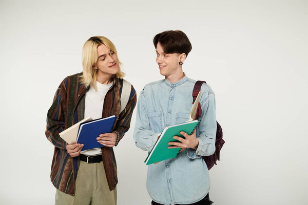 Two young people, part of the queer community, stand in a studio setting with notebooks in hand.