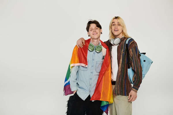 Two young people stand together, smiling and holding a pride flag, on a plain white background.
