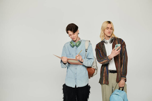 Two young queer students stand in casual attire against a grey background, one studying and the other on their phone.