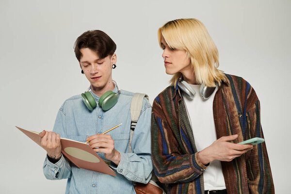 Two young queer students stand together in casual attire, one taking notes and the other looking on.