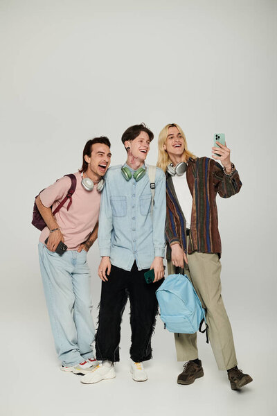 Three young men, dressed in casual attire, stand in a studio with a grey background and take a selfie.