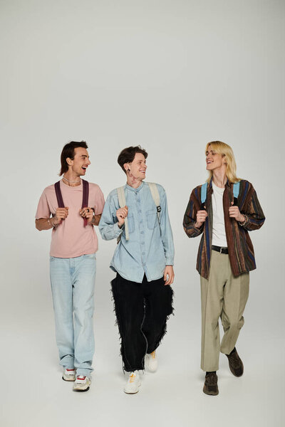 Three young students, wearing casual clothing and backpacks, walk side-by-side against a grey backdrop.