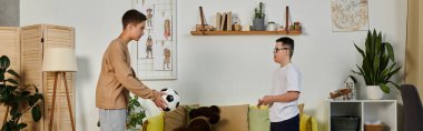 İki çocuk, biri futbol topu tutuyor, oturma odasında duruyor, arkadaşça bir oyun oynuyorlar..