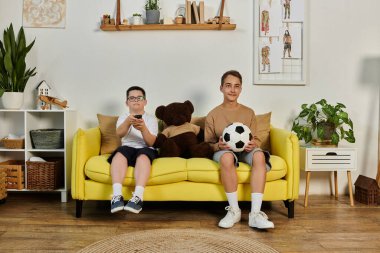 İki çocuk, biri Down sendromlu, sarı bir kanepede oturuyor..