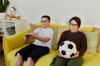 İki çocuk, biri futbol topu tutuyor, sarı bir kanepede rahatlıyor..