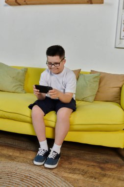 A young boy with Down syndrome smiles while playing on his phone on a yellow couch. clipart